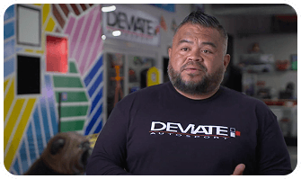 Man in black "DEVIATE Autostyling" shirt stands in a colorful, striped room with automotive decor.