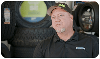 A man in a cap and polo shirt stands in front of stacked tires, with a blurred tire display in the background.