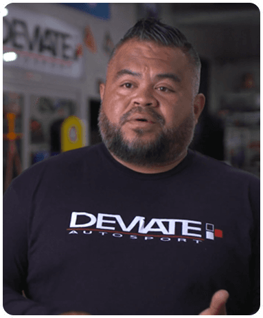 Man with a beard wearing a "DEVIATE AUTOSPORT" shirt, standing indoors with shelves and equipment in the background.