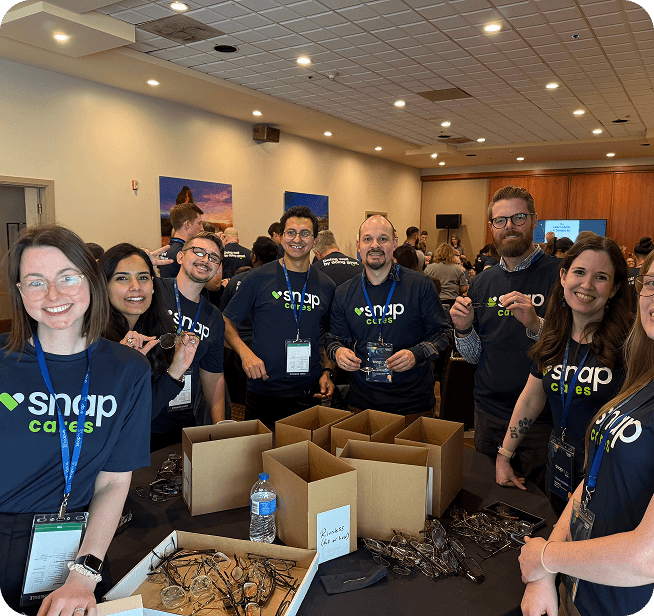 Group of people in matching "Snap Cares" shirts gathered around a table with boxes and supplies, smiling at an indoor event.
