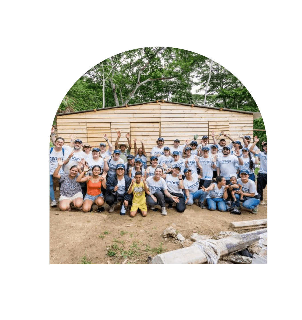 A large group of people wearing matching white t-shirts and caps are gathered in front of a partially constructed wooden building. They are smiling, and many are holding up peace signs with their hands. The setting appears to be outdoors in a wooded area.