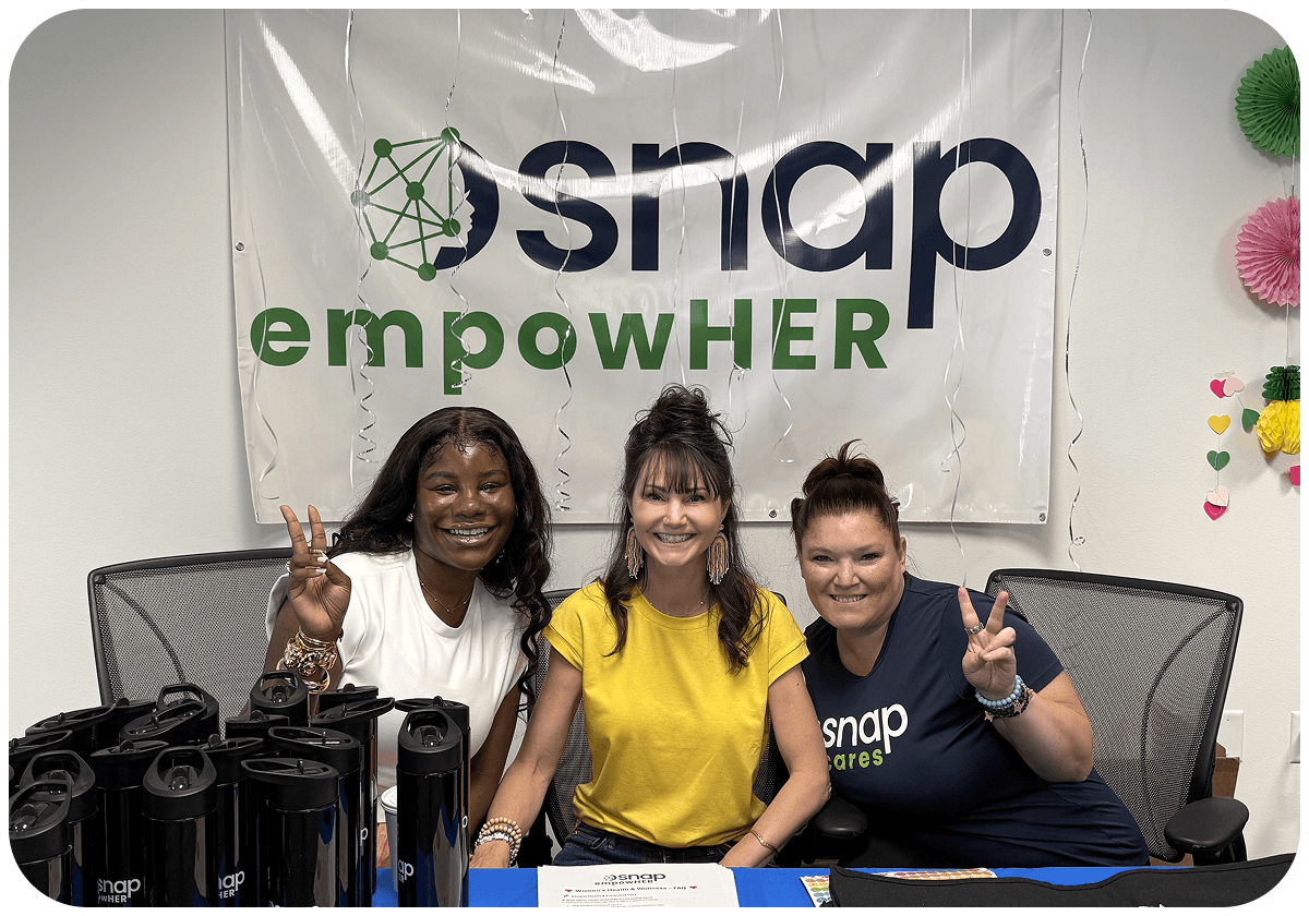 Three women smiling and making peace signs at a table with water bottles, in front of a "snap empowHER" banner.