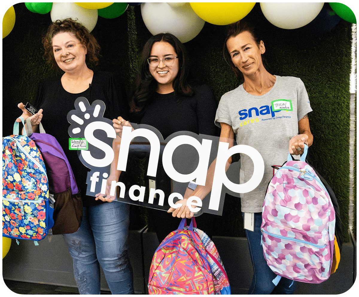 Three people holding colorful backpacks and a "snap finance" sign, standing in front of a wall with green and white balloons.