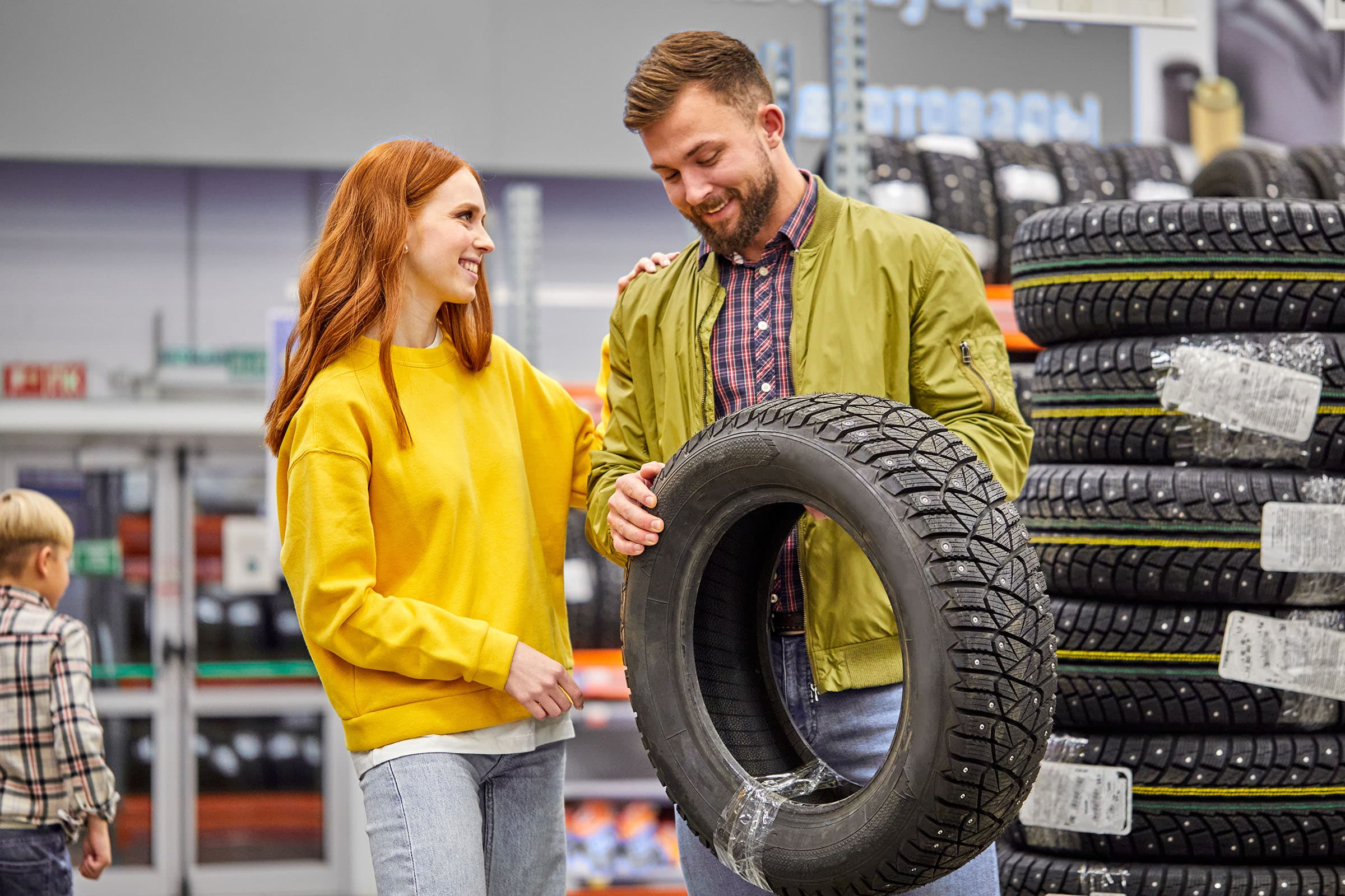 Smiling couple shopping for a tire in a store, with the man holding a winter tire and stacked tires visible in the background.