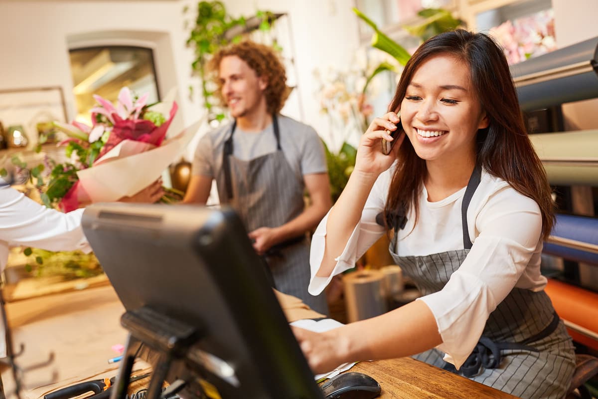 Smiling florist talking on the phone and using a computer at the counter, with another florist handing a bouquet to a customer.