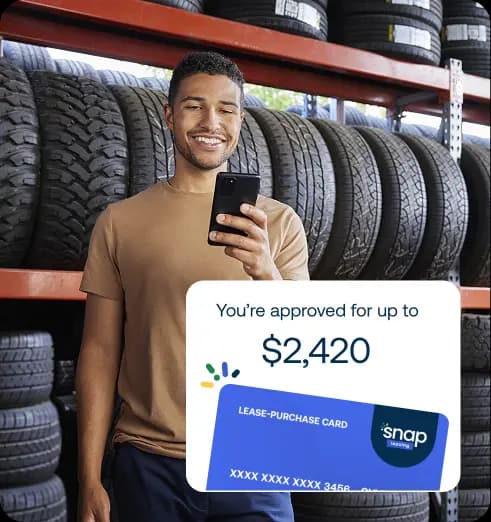 Man smiling while holding a phone in front of stacked tires. A digital card approval overlay shows "$2,420 lease-purchase card."