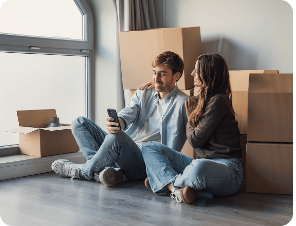 A couple sits on the floor of a new home, surrounded by moving boxes, smiling at a smartphone.