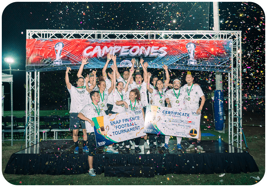 A soccer team celebrates on a podium, holding a "Champions" banner, certificates, and a trophy. Confetti fills the air under a "Campeones" sign.